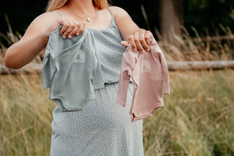 Babykleidung in Grau und Rosa, die von einer Frau im Freien gehalten wird, mit einem Waldhintergrund.