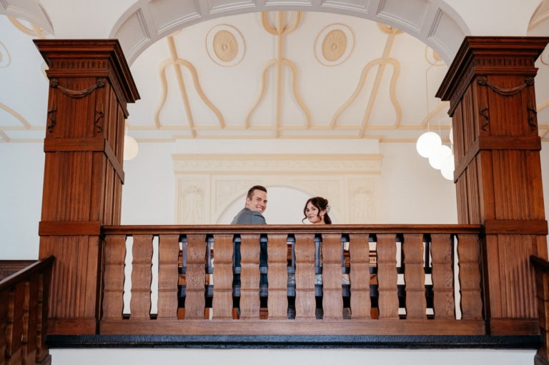 Hochzeitspaar auf Holzbalustrade, elegante Hochzeitsfotografie im stilvollen Saal, professionelle Hochzeitsfotos.