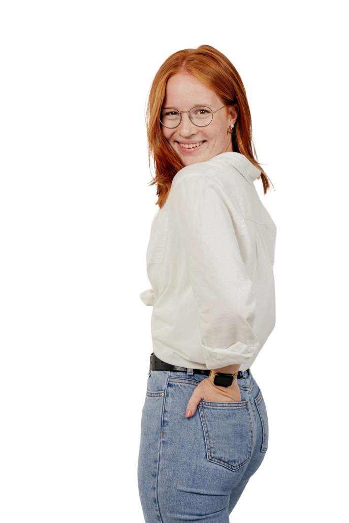 Fröhliche junge Frau mit roten Haaren, Brille und weißem Hemd, lächelnd und in Jeans, posiert vor weißem Hintergrund.