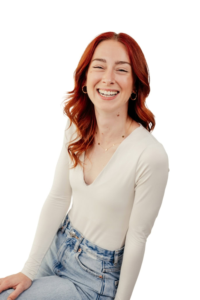 Eine lachende junge Frau mit roten Haaren, die in einem weißen Oberteil und Jeans gekleidet ist, sitzt vor einem weißen Hintergrund.