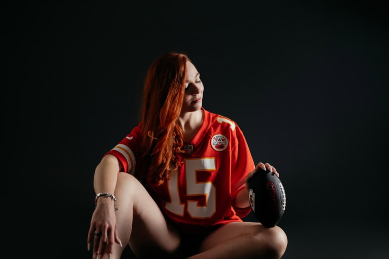 Eine junge Frau mit roten Haaren in einem roten Football-Trikot mit der Nummer 15, die auf einem dunklen Hintergrund sitzt und einen Football in der Hand hält.