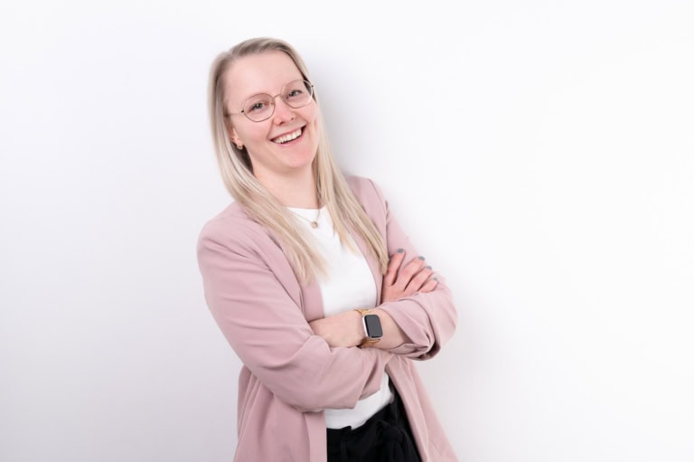 Fröhliche junge Frau mit Brille, blonden Haaren, rosa Blazer und weißen Hemd, lächelnd vor weißem Hintergrund.