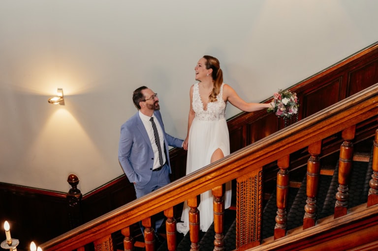 Braut und Bräutigam auf einer Treppe bei ihrer Hochzeit, lachend und glücklich, in eleganten Kleidern.