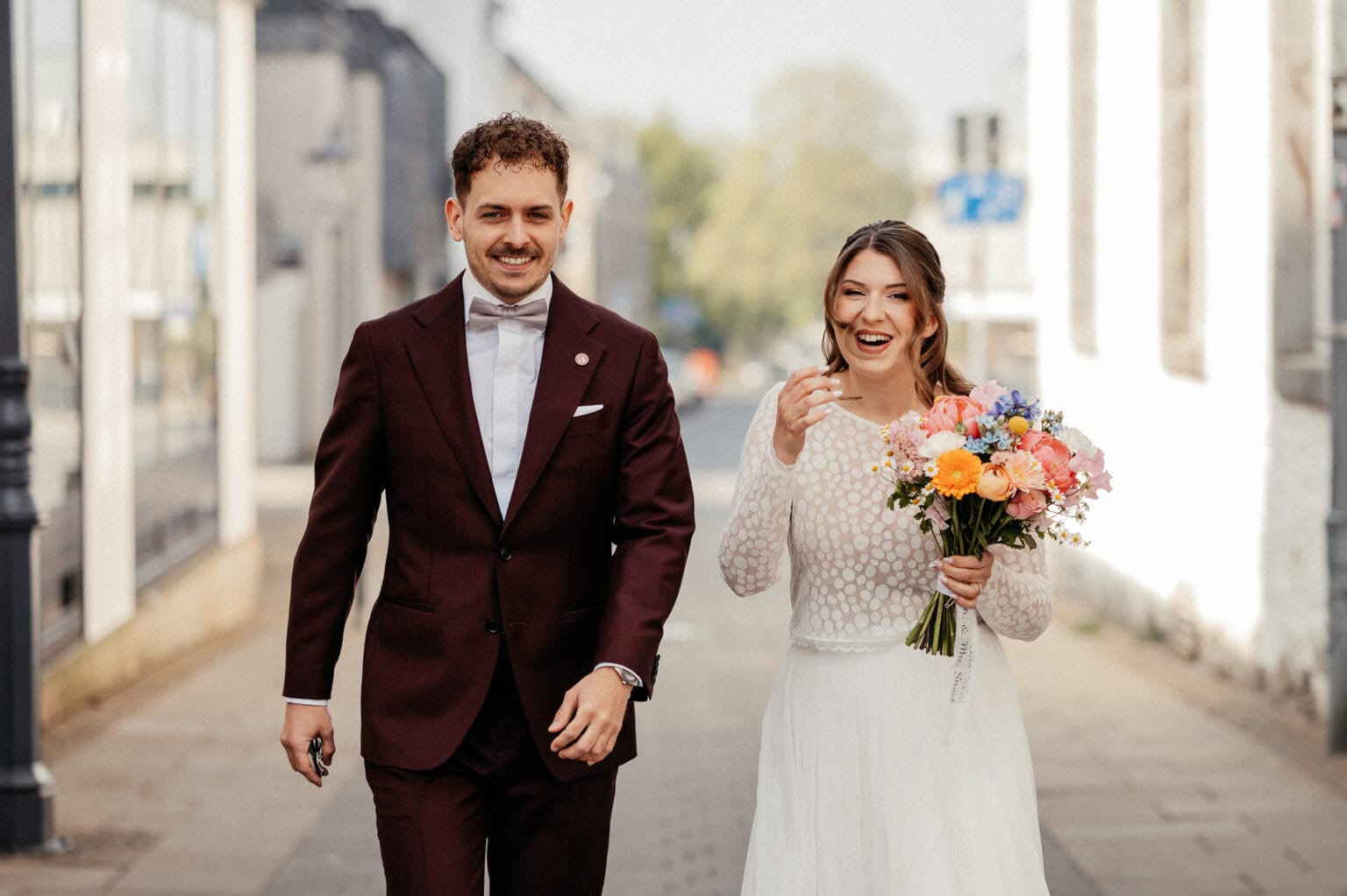 Fröhliches Brautpaar läuft Hand in Hand auf der Straße, die Braut hält einen bunten Blumenstrauß, beide lachen und genießen den Tag.