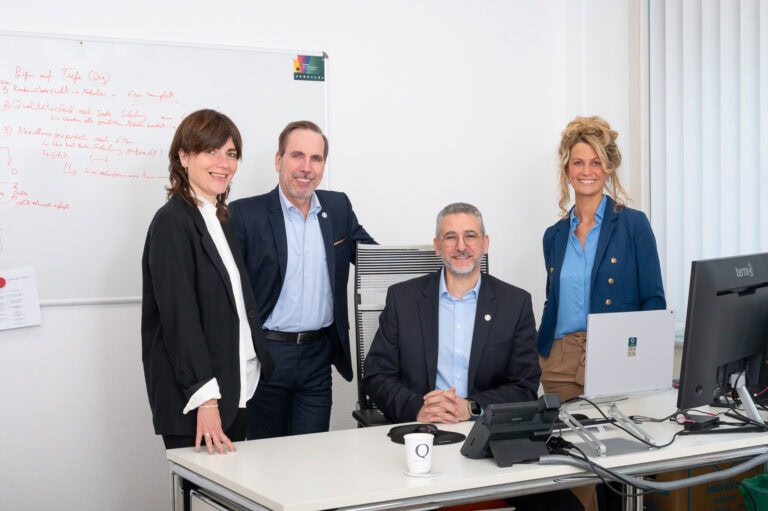 Vier Geschäftsleute in einem modernen Büro, lachen und posieren für das Foto, mit Computer und Whiteboard im Hintergrund.