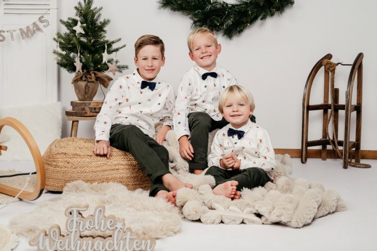 Weihnachtskinder sitzen auf einem flauschigen Teppich vor einem festlich geschmückten Hintergrund mit Weihnachtsbaum und Dekorationen.