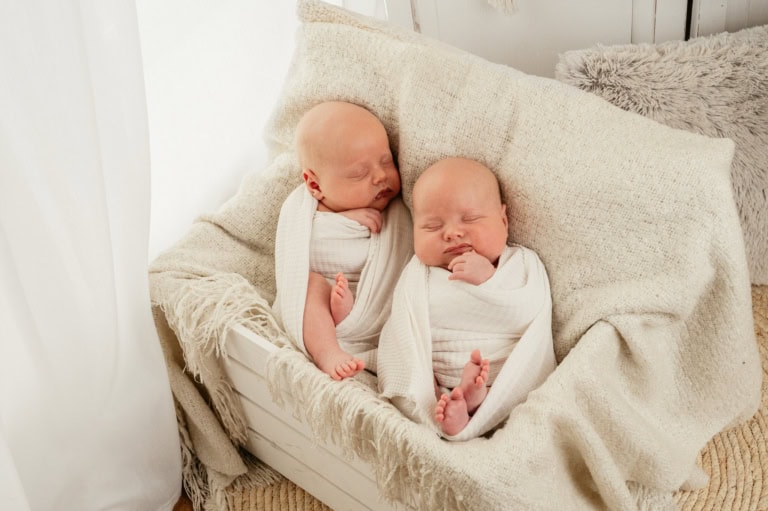 Alt Text: Zwillingsbabys schlafen friedlich auf einem Bett, eingewickelt in weiße Decken, umgeben von kuscheligen Kissen.