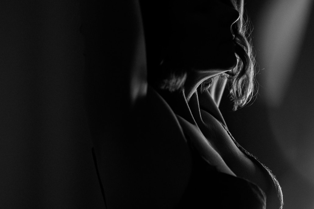Eine dunkle Silhouette einer Frau mit welligem Haar, die im Schatten steht und nur teilweise sichtbar ist. Die Bildkomposition betont die Konturen und das Spiel von Licht und Schatten.