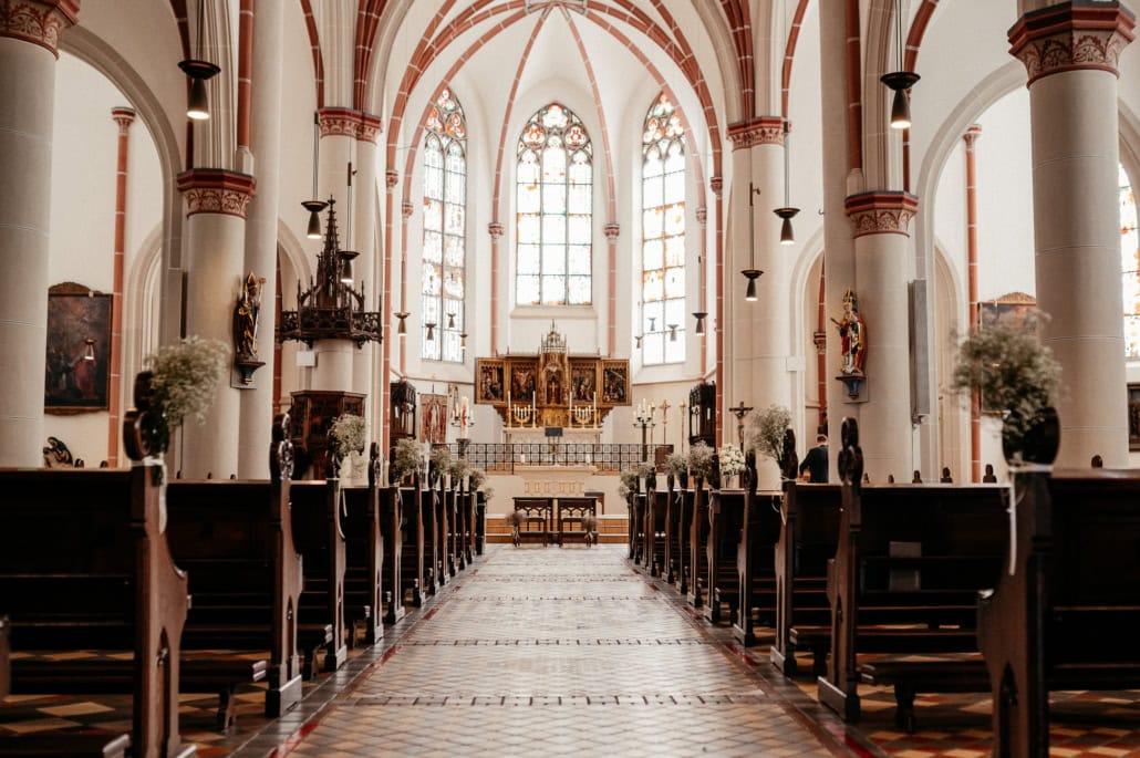 Der Innenraum einer Kirche mit hohen, bunten Fenster und Altar im Hintergrund, umgeben von Säulen und Kirchenbänken, mit wenigen Personen im Gebet.