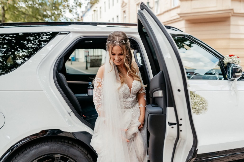 Glückliche Braut in Hochzeitskleid beim Ausstieg aus einem weißen Luxusauto, im urbanen Umfeld mit Bäumen und Gebäuden im Hintergrund.