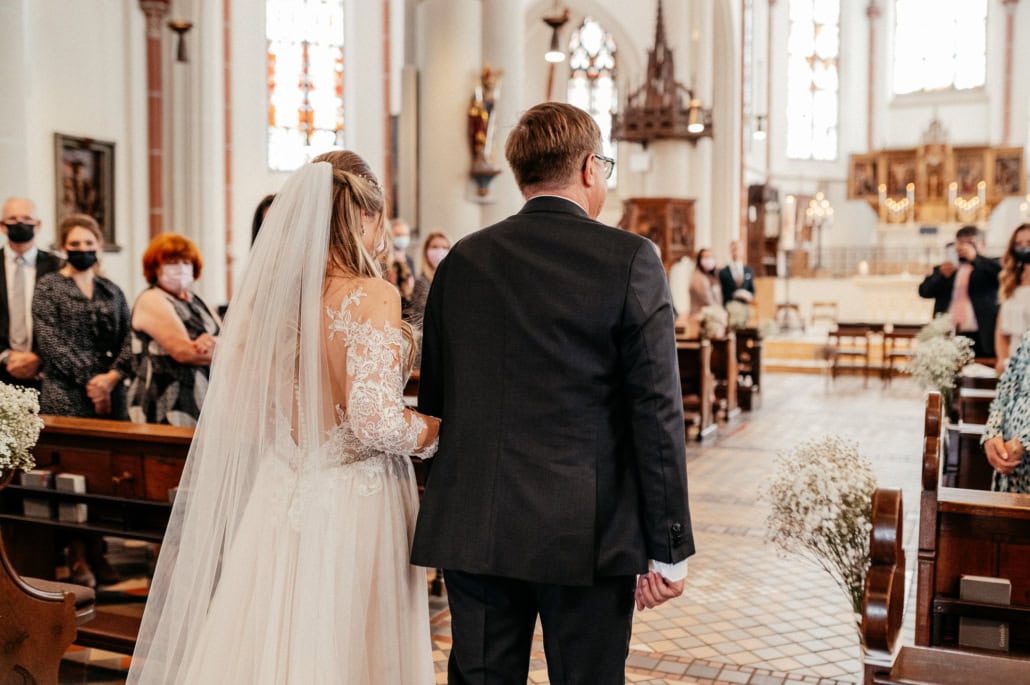 Hochzeitspaar bei der Trauung in einer Kirche, Braut in weißem Kleid mit Schleier, Bräutigam in dunklem Anzug, umgeben von Gästen, die Masken tragen, festliche Atmosphäre, helle Fenster und religiöse.