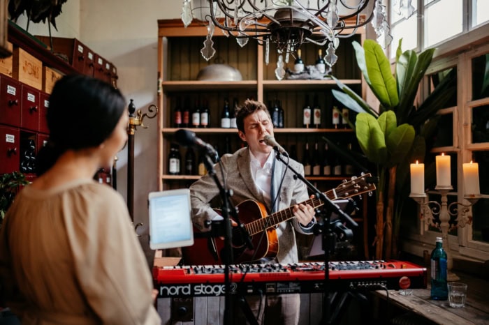 Ein Mann spielt Gitarre und singt in einem gemütlichen Raum mit Kerzen und Pflanzen, während eine Frau ihm zuhört.