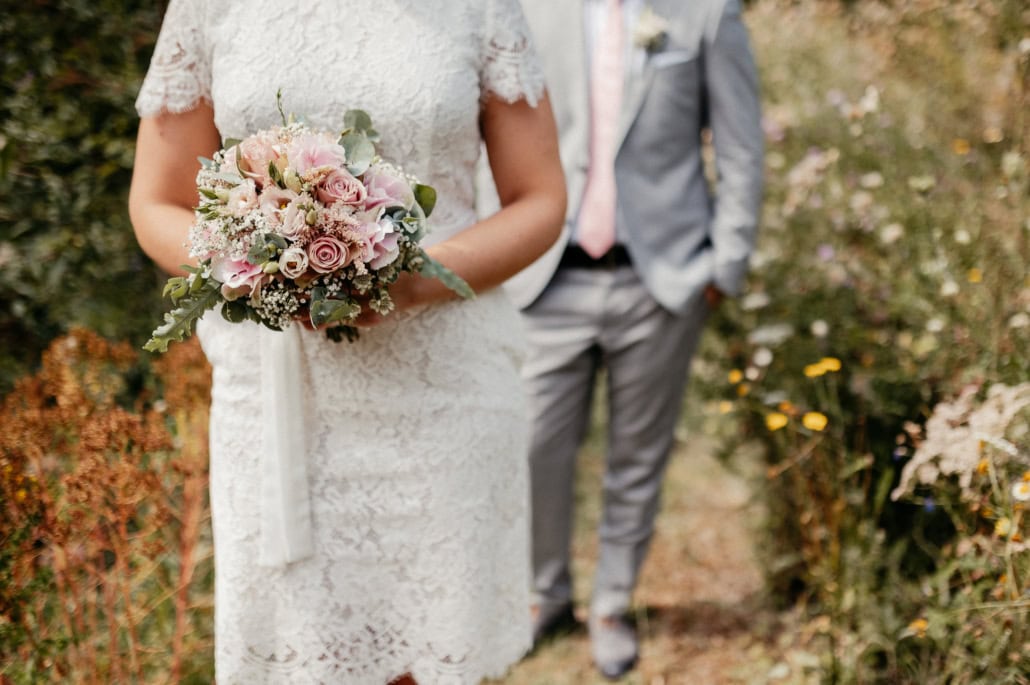 Blumenstrauß mit rosa und weißen Blumen, gehalten von einer Braut in einem weißen Spitzenkleid bei einer Hochzeit im Freien.