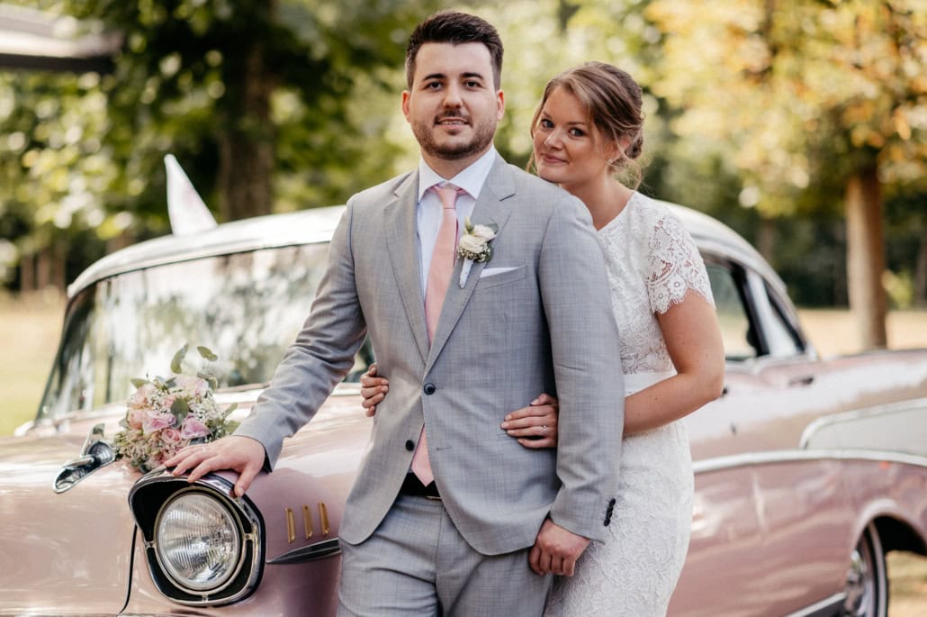 Glückliches Brautpaar vor einem Vintage-Auto im Park, bei ihrer Hochzeit im Freien, umgeben von Herbstbäumen.
