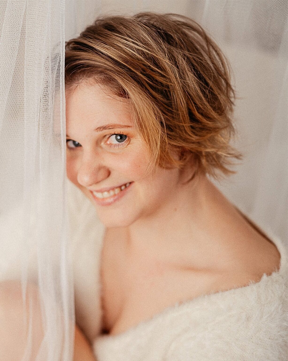 Nahaufnahme einer lachenden jungen Frau mit kurzen, lockigen, hellbraunen Haaren, die hinter einem transparenten Vorhang steht.