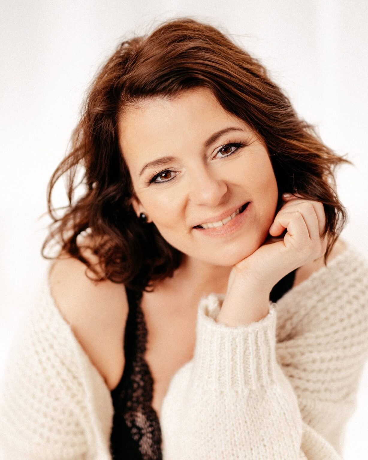 Freundliche junge Frau mit braunen, lockigen Haaren, lächelnd und in einem weißen Pullover, vor hellem Hintergrund.