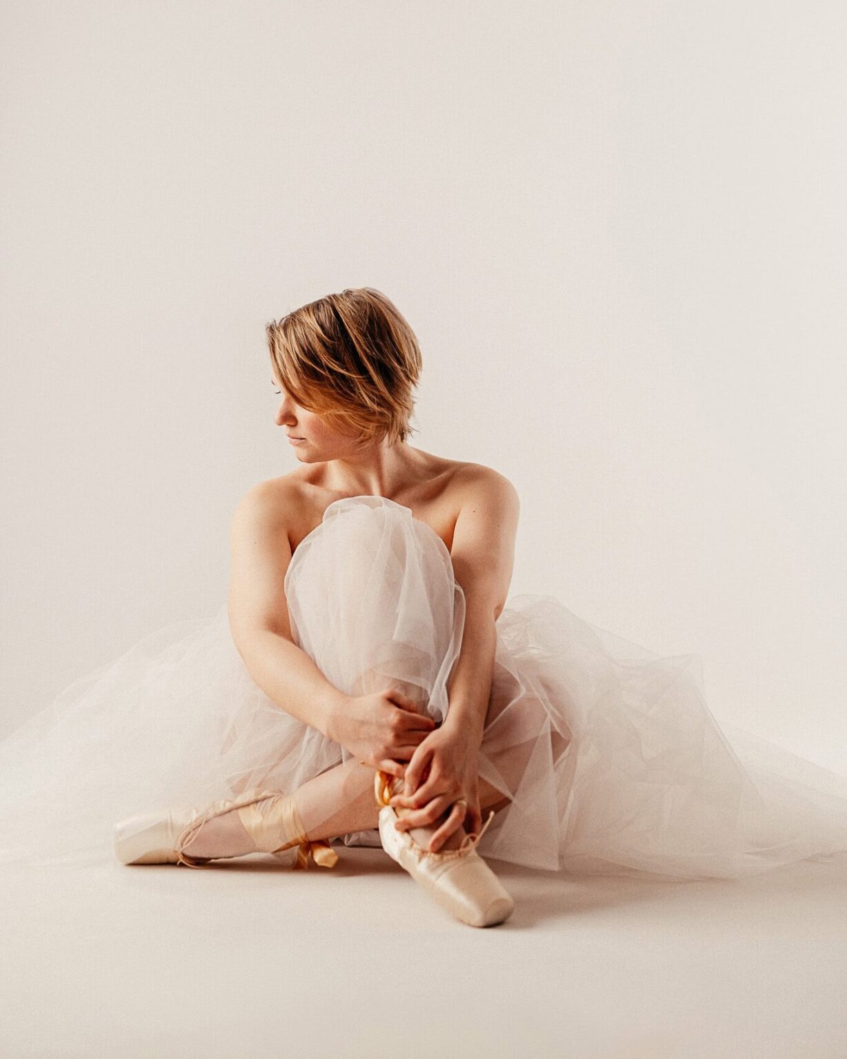 Balletttänzerin in weißem Tutu sitzt auf dem Boden, die Beine angezogen, vor einem weißen Hintergrund, nachdenklich und ruhig wirkend.
