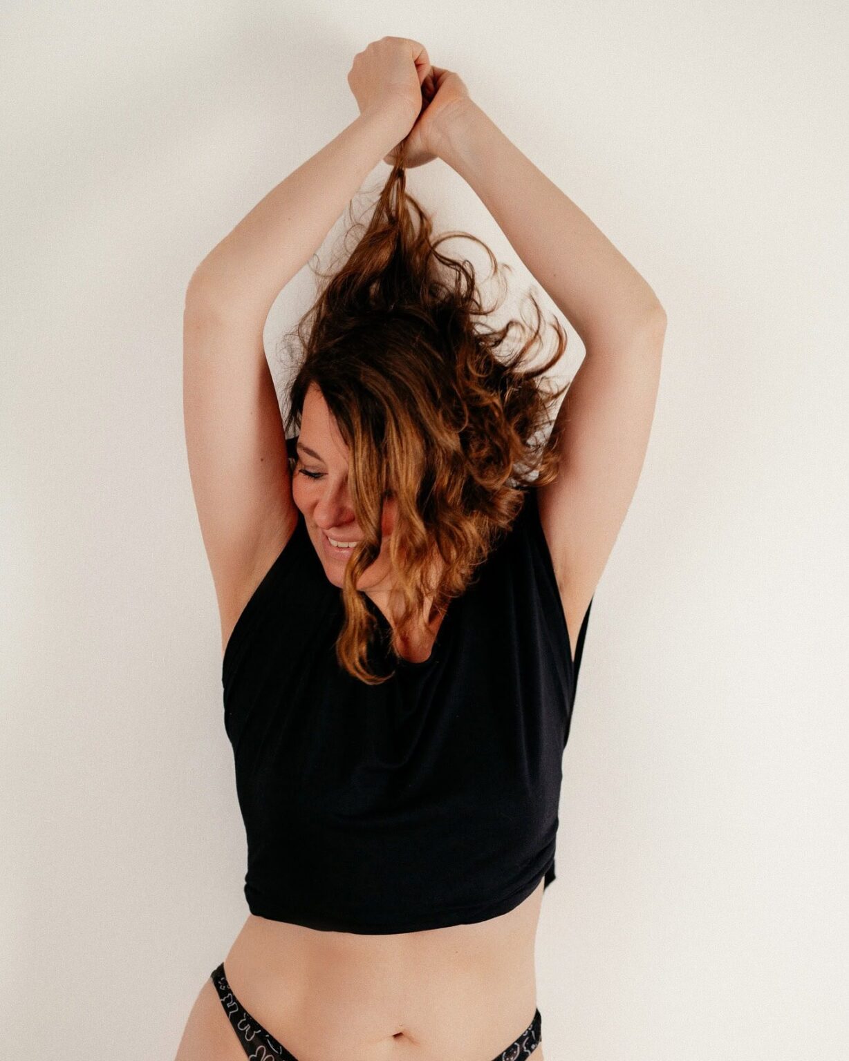 Junge Frau mit lockigem Haar, die sich freut und die Arme über dem Kopf hebt, vor hellem Hintergrund.