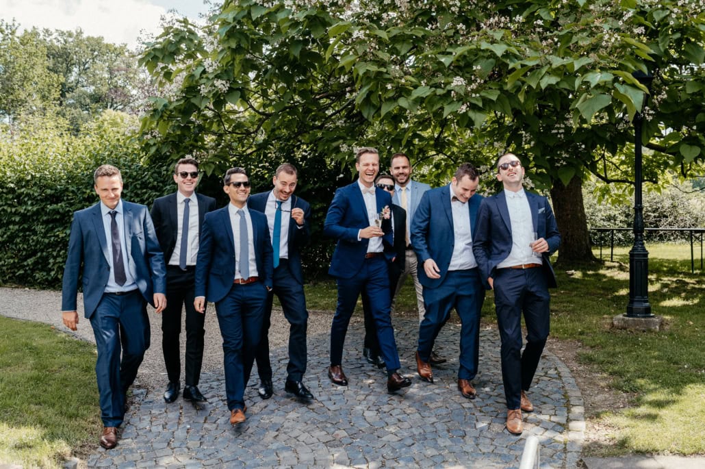Gruppe von acht Männern in Anzügen beim Spaziergang im Park, lachend und entspannt.