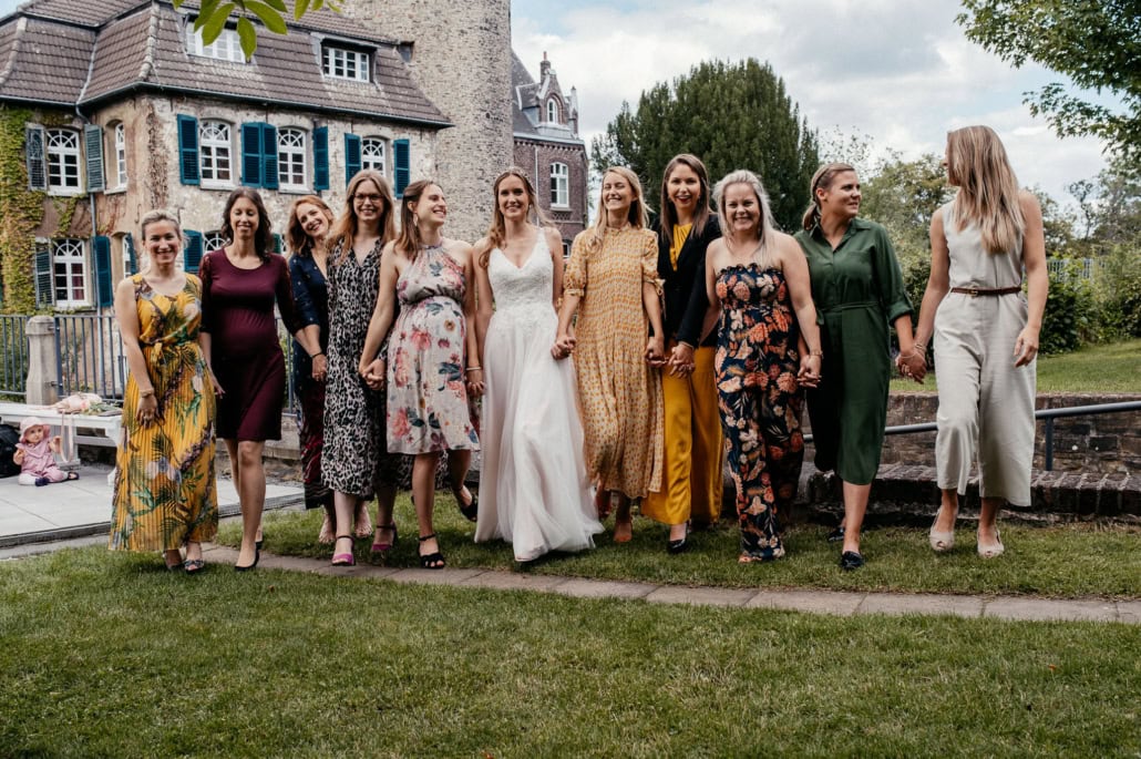 Gruppe von Frauen in eleganten Kleidern beim Spaziergang im Garten vor einem historischen Gebäude.